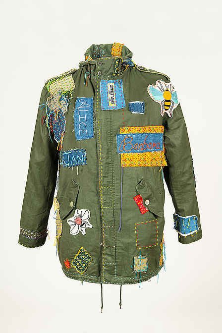Eine olivgrüne Outdoorjacke ist mit diversen auffälligen farbenfrohen Patches repariert: Ins Auge stechen zwei Bügelflicken mit Insektenmotiven, außerdem großflächig gestickte Namen auf Bügelflicken, die auf die Beteiligten des Berliner Kollektivs "STREETWARE saved item" hinweisen. 