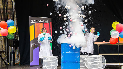 Aus einem blauen Fass explodieret eine weißer Nebel mit bunten Luftballons.