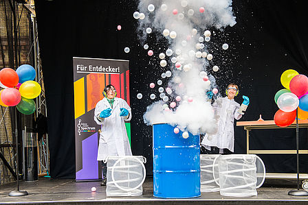 Aus einem blauen Fass explodieret eine weißer Nebel mit bunten Luftballons.