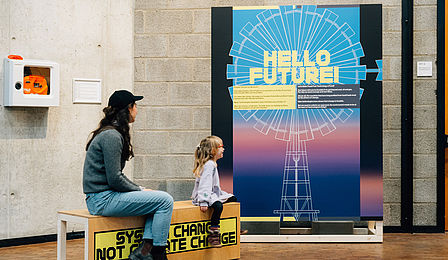 Eine Frau und ein kleines Mädchen sitzen auf einer Bank, die mit dem Slogan "System change, not climate change" beschriftet ist. Auf einem Plakat steht "Hello Future".