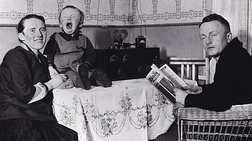 100 Jahre Radio - eine historische Schwar-Weisß-Aufnahme zeitgt eine Familie vor einem Radioempfänger. Mutter und Vater posieren für das Foto, das Kleinkind weint.