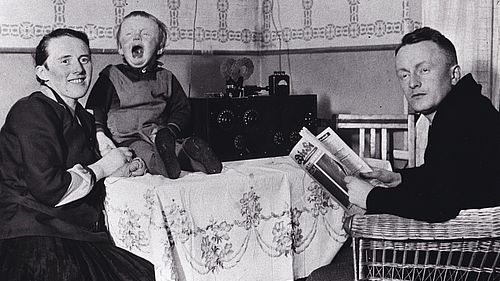 100 Jahre Radio - eine historische Schwar-Weisß-Aufnahme zeitgt eine Familie vor einem Radioempfänger. Mutter und Vater posieren für das Foto, das Kleinkind weint.
