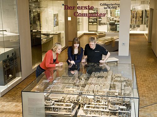 Zwei Frauen und ein Mann beugen sich über eine große Glasvitrine, in der das mechanische Rechenwerk der Z1 zu sehen ist.