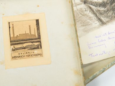 Aufgeklappte Schachtel, darin rechts eine Zeichnung mit einer Widmung. Links ist die Deckelinnenseite zu sehen, auf der Heinrich Hirschbergs Exlibris klebt. Es zeigt im Vordergrund ein aufgeschlagenes Buch auf einem Tisch, im Hintergrund ist durch einen Fensterrahmen eine Fabrik zu erkennen.