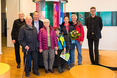 Man sieht eine Gruppe von Menschen mit einem Blumenstrauß vor einem Raum, dessen Wände bunt beklebt sind.