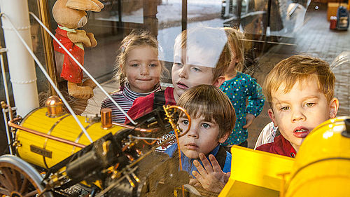 Fünf Kinder gucken in eine Vitrine, in der sich das Modell einer gelben Lokomotive befindet. Auf der linken Seite ist eine Hasen-Stoffpuppe zu sehen.