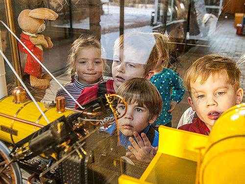 Fünf Kinder gucken in eine Vitrine, in der sich das Modell einer gelben Lokomotive befindet. Auf der linken Seite ist eine Hasen-Stoffpuppe zu sehen.