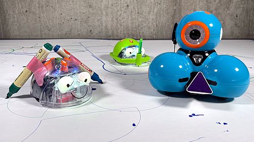 Drei Roboter mit montierten Stiften stehen auf weißem Untergrund. Auf dem Untergrund sind geometrische Formen gezeichnet.