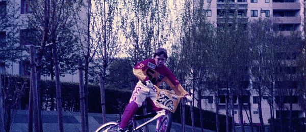 A child rides a BMX bike against a backdrop of highrises. He is braking and skidding sideways. The child is wearing BMX shorts and a shirt, but no helmet, and attached to the handlebars of his bike is a race plate for use in competitions.