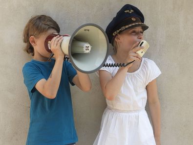 Zwei Kinder stehen nebeneinander. Der Junge versucht so durch ein Megaphon zu schauen als wäre es ein Fernrohr. Das Mädchen trägt eine blaue Zugschaffnermütze und spricht in den Lautsprecher des Megaphons, der durch ein Kabel mit dem Gerät verbunden ist.