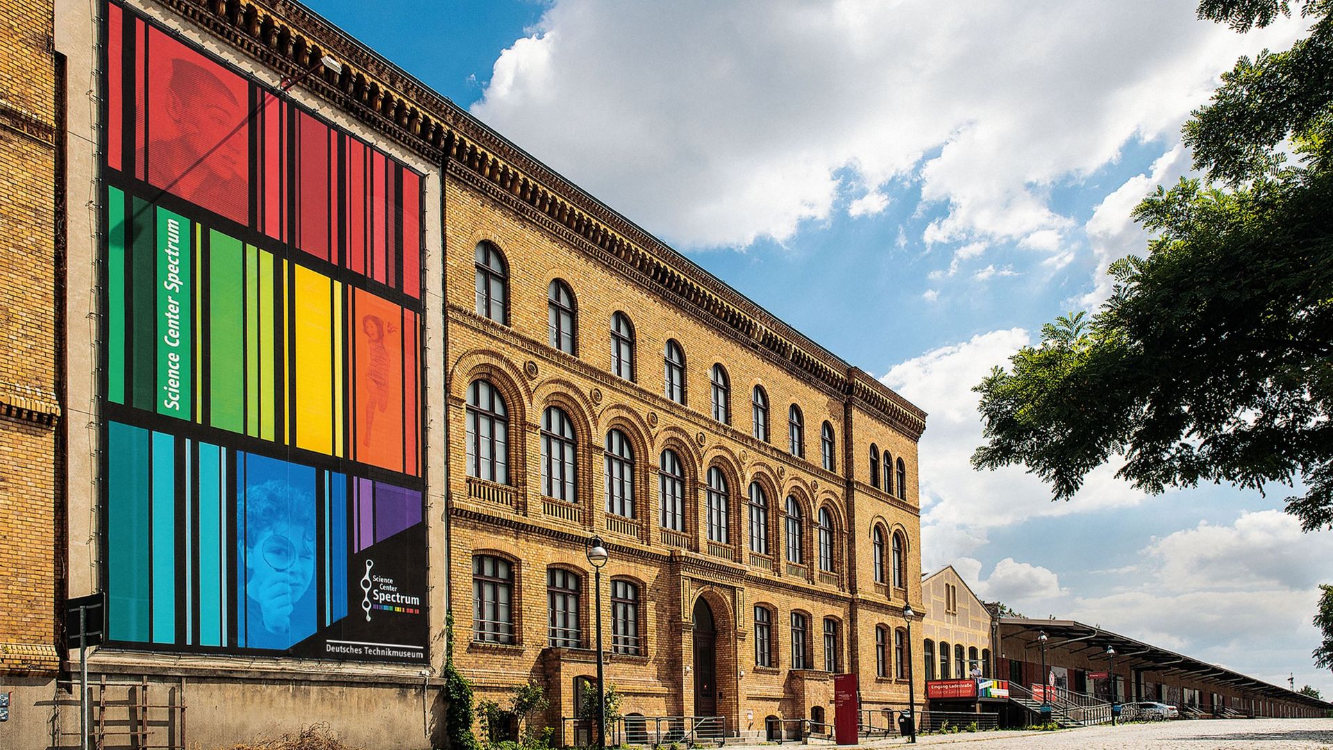 Ein dreigeschossiger Backsteinbau mit einem bunt gestreiften Transparent an der Fassade. Davor eine kopfsteingepflasterte Straße.