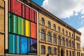 Ein dreigeschossiger Backsteinbau mit einem bunt gestreiften Transparent an der Fassade. Davor eine kopfsteingepflasterte Straße.