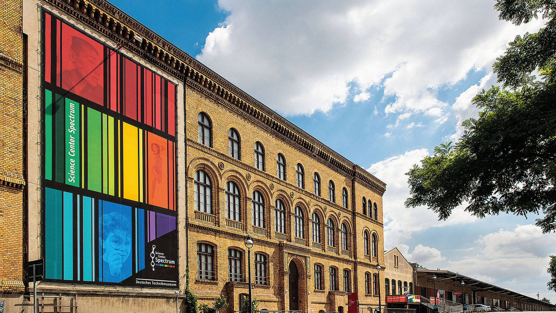 Ein dreigeschossiger Backsteinbau mit einem bunt gestreiften Transparent an der Fassade. Davor eine kopfsteingepflasterte Straße.