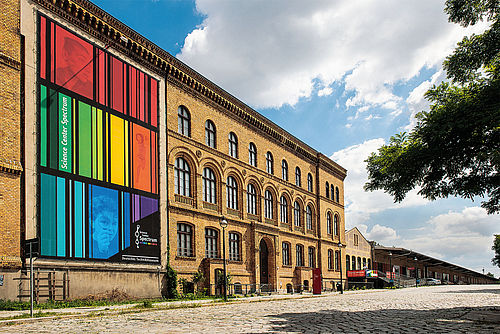 Ein dreigeschossiger Backsteinbau mit einem bunt gestreiften Transparent an der Fassade. Davor eine kopfsteingepflasterte Straße.