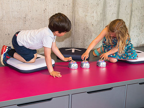 Zwei Kinder sitzen auf Kissen auf einem rosa Untergrund. Sie beugen sich nach vorne und programmieren die Roboter Blue-Bot.