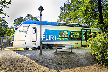 Seitenansicht von einem blau-weißen Zug mit der Aufschrift "FLIRT Akku". Der Zug steht auf einem Bahnsteig im Grünen ein, davor eine Laterne und eine Bank.