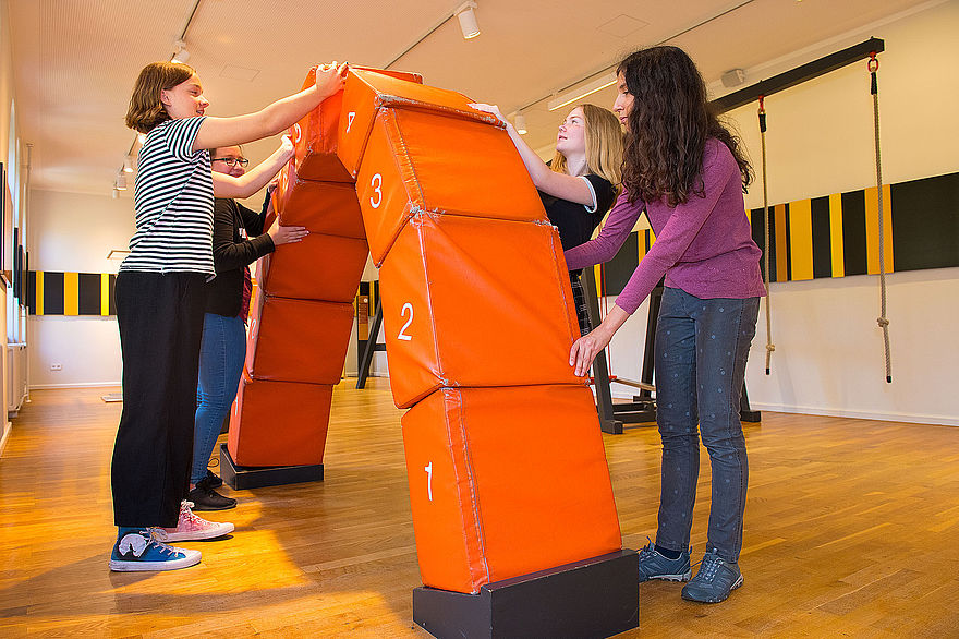In einem hellen Raum steht ein aus orangenen Blöcken zusammengesetzter Brückenbogen. Auf den einzelnen Blöcken befinden sich Zahlen. Auf den untersten eine 1, auf den nächsten eine 2, auf dem höchsten ein 5. Der Brückenbogen ist etwa so groß wie die vier Mädchen, die vor und hinter dem Bogen stehen. Sie halten einzelne Elemente mit den Händen.