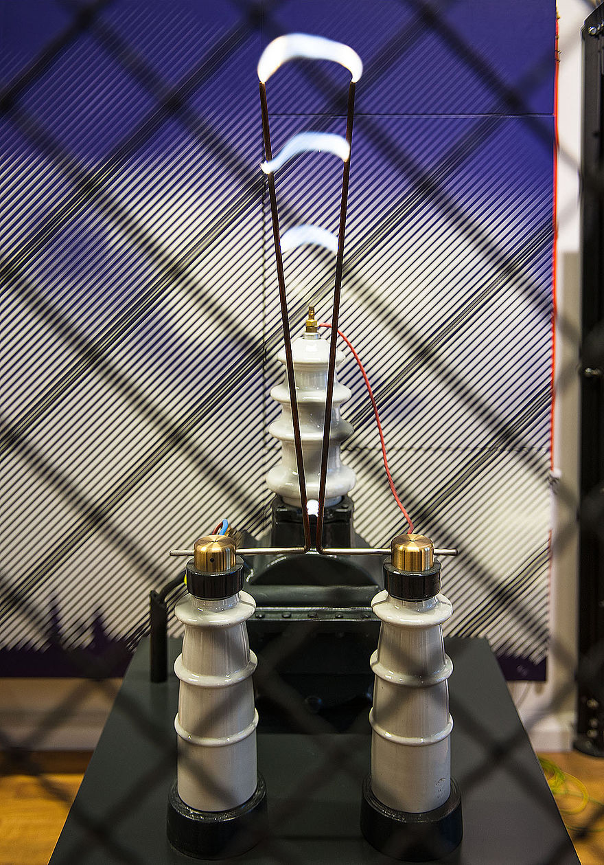 Hinter einem Zaun befindet sich ein Experiment. Auf einem dunklen Sockel stehen vorn zwei weiße Säulen, deren Spitzen metallisch sind. Darüber ragen zwei dünne schwarze Stäbe wie ein V in die Luft. Zwischen ihnen spannen sich Lichtbögen. Hinter den Stäben ist eine weitere weiße Säule zu erkennen.