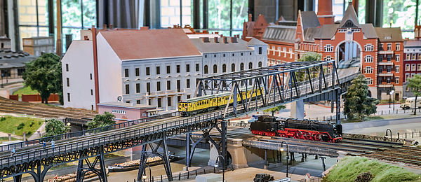 Ein detailreiches Eisenbahnmodell: Eine Hochbahnbrücke, auf der eine gelbe Bahn fährt, kreuzt Eisenbahnschienen, einen Kanal und eine Straße. Im Umfeld Modellhäuser und Miniaturen von Autos, Loks und Schiffen.