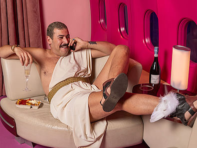 Das Farbfoto zeigt einen Mann mit Schnurrbart. Er liegt entspannt in einer luxuriösen Umgebung, die an das Innere eines Flugzeugs erinnert. Er trägt eine Art Toga mit goldenem Gürtel, an den Füßen Sandalen mit Flügeln. In einer Hand hält er ein Glas Sekt, in der anderen ein Telefon. Neben ihm stehen eine Pappschale mit Pommes Frites. Auf einem Sofa ihm gegenüber liegt ein Helm mit Flügeln. Die Szenerie ist in kräftigem Pink gehalten.