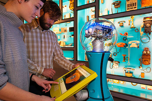 Eine Frau und ein Mann stehen mit Museumsroboter TIM vor einer Vitrine. Gerade betätigt sie den Touchbildschirm des Roboters.