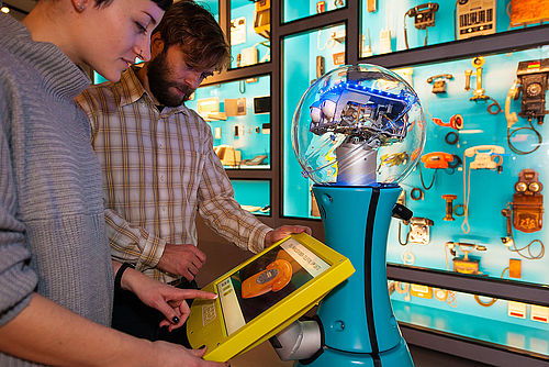 Eine Frau und ein Mann stehen mit Museumsroboter TIM vor einer Vitrine. Gerade betätigt sie den Touchbildschirm des Roboters.
