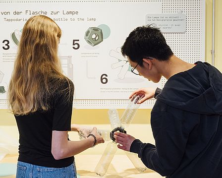 Vor einer Text- und Bildtafel mit der Aufschrift „von der Flasche zur Lampe“ schrauben ein Mädchen und ein Junge gemeinsam leere Plastikflaschen in eine Kunststoff-Fassung. Das Ergebnis ist eine lustig geformte Lampe.