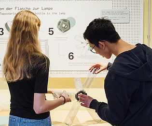 Vor einer Text- und Bildtafel mit der Aufschrift „von der Flasche zur Lampe“ schrauben ein Mädchen und ein Junge gemeinsam leere Plastikflaschen in eine Kunststoff-Fassung. Das Ergebnis ist eine lustig geformte Lampe.