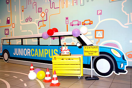 Ein etwa sechs Meter langer Aufsteller aus Pappe in Form eines Autos. Auf der Seite ist der Schriftzug „Junior Campus“ angebracht. Er ist mit Luftballons geschmückt. Davor steht ein Werkzeugschrank und ein Verkehrsschild mit der Aufschrift „Achtung Geburtstagsfeier“.
