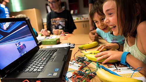 Kinder sitzen an einem Tisch vor einem Laptop. Auf dem Bildschirm ist ein Spiel mit einem Rennauto zu sehen. Die Kinder steuern das Spiel mit Bananen, die auf dem Tisch liegen und über Kabel und ein Gerät mit dem Laptop verbunden sind.