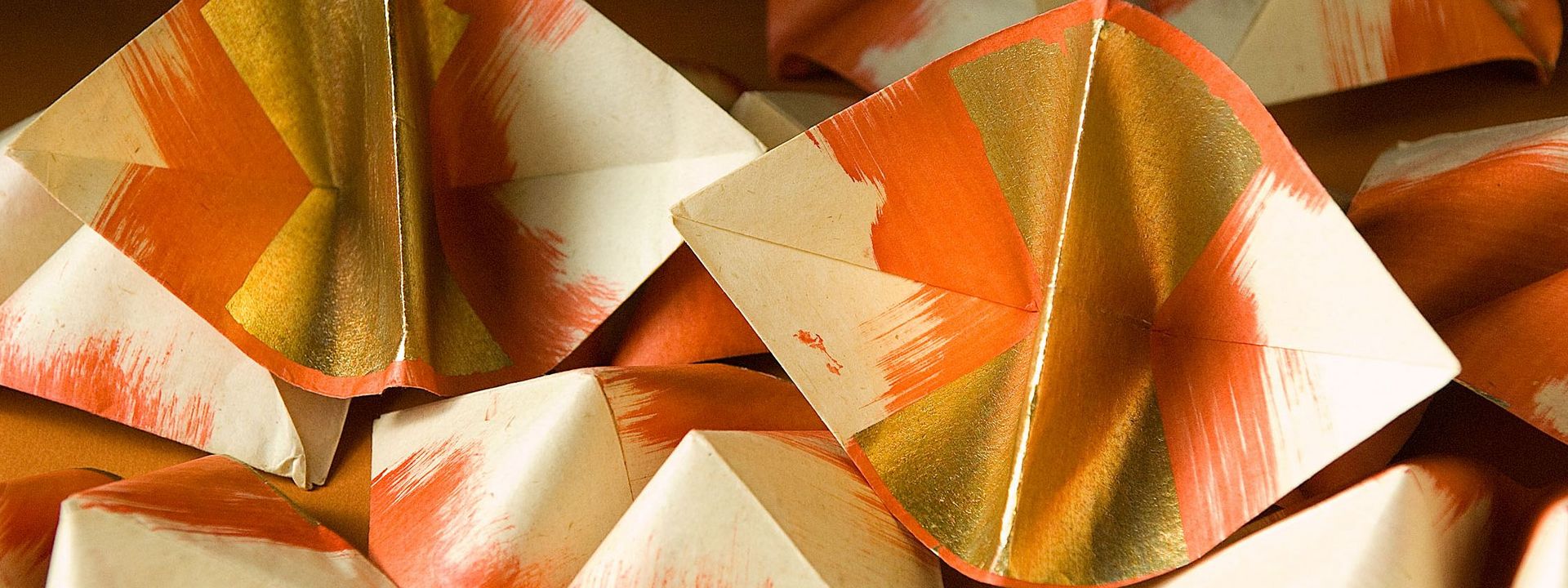 Eine Vielzahl gefalteter Papierschiffchen. Das Papier der Schiffchen verläuft von den entgegenliegenden Außenkanten zur Mitte von weiß nach rot. Die Faltmitte ist mit Goldblatt veredelt.