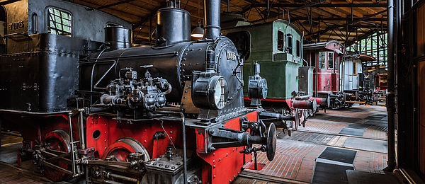 Blick in die Schienenverkehrs-Ausstellung: Eine historische schwarzrote Lokomotive, und mehrere Abteilwagen stehen auf Schienen nebeneinander aufgereiht.