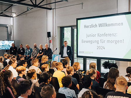 Ein Koferenz-Raum, an der Wand ein großer Bildschrim mit der Aufschrift "Hertlich willkommen, Junior Konferenz: Bewegung für morgen! 2024", die Stuhlreihen sind besetzt mit Schülerinnen und Schülern, am Stehpult steht ein Mann im Anzug und begrüßt die Kinder, links daneben sitzen sechs Erwachsene auf Stehhockern.