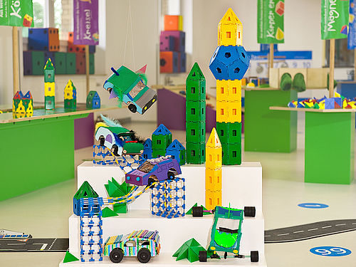 In einem Raum des Junior Campus stehen kleine selbstgebaute Fahrzeuge aus Pappe und Türme, die aus geometrischen Flächen aus Plastik zusammengesteckt wurden, auf einem dreistufigen Podest. Im Hintergrund befinden sich Arbeitstische mit weiteren Materialien.