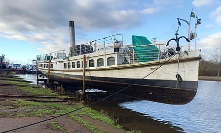 Mansieht ein Schiff am Ufer eines Gewässers, auf einem Gestell vertäut.