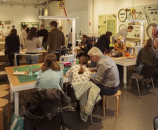 Blick in die Ausstellungen "Reparieren" des Deutschen Technikmuseums. Einige Menschen sitzen um Tische und sind gemeinsam etwas am reparieren.