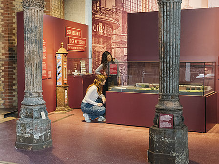 Zu sehen ist die in weinrot gehaltene Themeninsel „Eisenbahn in der Metropole“ der überarbeiteten Ausstellung Schienenverkehr im Deutschen Technikmuseum. Im Vordergrund zwei alte, verzierte Metallsäulen, die sogenannten Hartungschen Säulen. Im Hintergrund betrachten zwei Personen den Inhalt einer Vitrine.