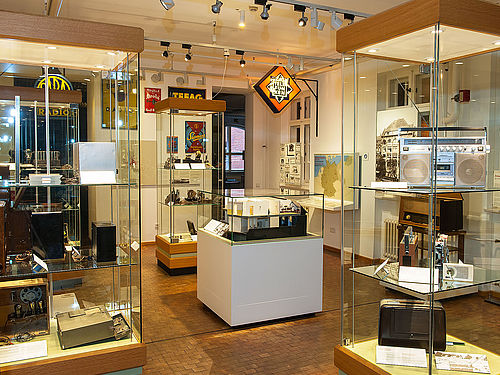 Mehrere Vitrinen in der Ausstellung zur Nachrichtentechnik. In den Vitrinen befinden sich auf mehreren Ebenen verschiedene Radiogeräte und Empfänger. Im Zentrum steht das Modell eines historischen Fernsehstudios, darüber hängt ein beleuchtetes Werbeschild.