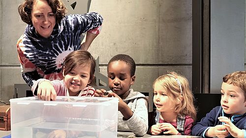 Vier Kinder sitzen um einen Tisch. Eine Museumspädagogin steht hinter der Gruppe. Auf dem Tisch steht eine durchsichtige, viereckige Wanne mit Wasser. Ein Mädchen setzt vorsichtig setzt vorsichtig einen Gegenstand auf das Wasser. Die Kinder beobachten interessiert was geschieht.