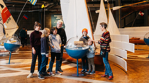 Vier Kinder und drei erwachsene Personen befinden sich in der Schifffahrtsausstellung. Eine Mitarbeiterin des Museum erklärt an einer bojenähnlichen Vitrine ein Objekt. 