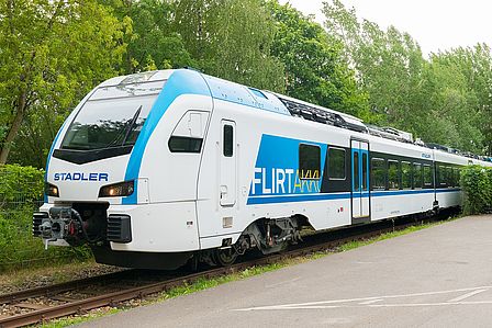 Dreiviertelansicht eines blau-weißen Zugs mit den Aufschriften „Stadler“ und „FLIRT Akku“. Der Zug fährt auf einem Gleis im Grünen, davor ist eine asphaltierte Fläche zu sehen.