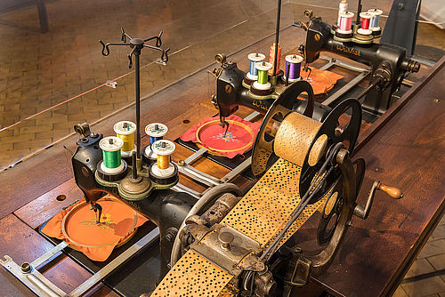 Blick von oben auf eine große, historische Stickmaschine mit einer Spule mit aufgerollter Lockkarte und drei Stickköpfen mit verschiedenfarbigem Garn und eingespannten Stickrahmen.