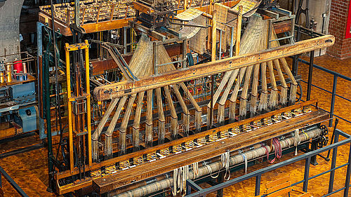 Ein großer, historischer Bandwebstuhl aus Holz und Metall. Auf der Oberseite des Webstuhls sind zwei Jacquardaufsätze mit Lochkarten zu erkennen, mit denen das Muster des späteren Stoffs vorgegeben wird.