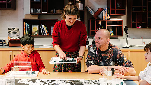 Zwei Erwachsene und zwei Kinder sitzen an einem Tisch. Vor ihnen sind verschiedene Workshopmaterialien aufgebaut, wie Eiswürfel, Wasserbehälter und blauer Farbstoff.