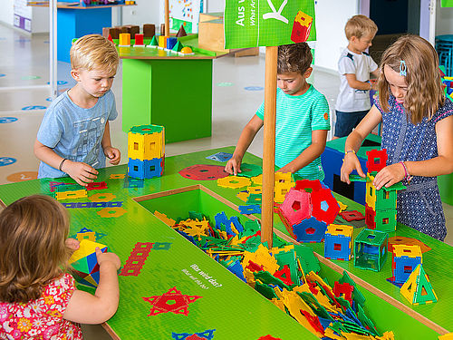 Vier Kinder stehen um einen bunten Arbeitstisch, in dessen Mitte in einer Vertiefung geometrische Formen aus buntem Kunststoff liegen. Sie stecken die Formen zu Würfeln, Türmen und Pyramiden zusammen. 