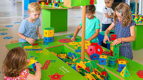 Vier Kinder stehen um einen bunten Arbeitstisch, in dessen Mitte in einer Vertiefung geometrische Formen aus buntem Kunststoff liegen. Sie stecken die Formen zu Würfeln, Türmen und Pyramiden zusammen. 
