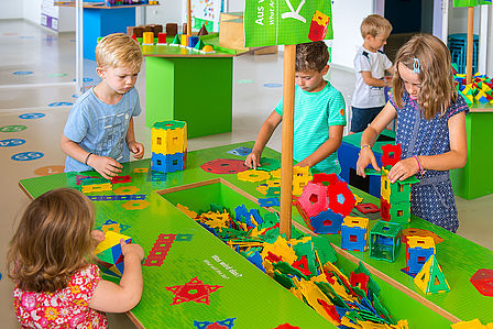 Vier Kinder stehen um einen bunten Arbeitstisch, in dessen Mitte in einer Vertiefung geometrische Formen aus buntem Kunststoff liegen. Sie stecken die Formen zu Würfeln, Türmen und Pyramiden zusammen. 