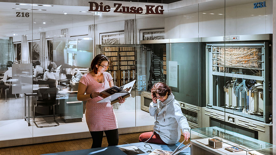 Blick in die Informatik-Ausstellung: Eine Frau sitzt auf einem Podest und hält sich den Hörer einer Hörstation ans Ohr. Eine andere Frau steht neben ihr und liest in einem großen Heft. In wandhohen Vitrinen im Hintergrund sind historische Rechenmaschinen zu sehen. Von der Decke hängt der Schriftzug „Die Zuse KG“.
