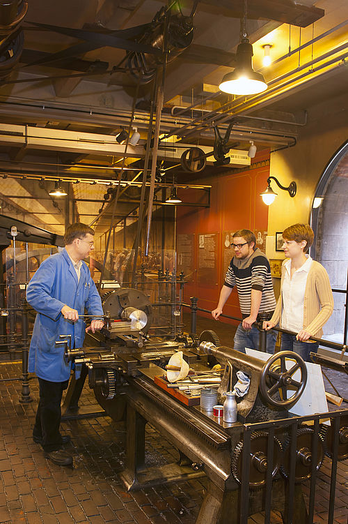 Ein Vorführer im blauen Arbeitskittel demonstriert zwei Museumsgästen eine historische Werkbank. Sie hat verschiedene Zahnräder, Kurbeln und Handräder. In die Maschine ist ein Metallstück eingespannt.