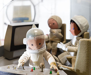 A Sandman wearing a cosmonaut’s suit is standing at a control panel with levers. In the background there are two more wooden puppets sitting in a space station beneath a glass roof with satellite dishes. 